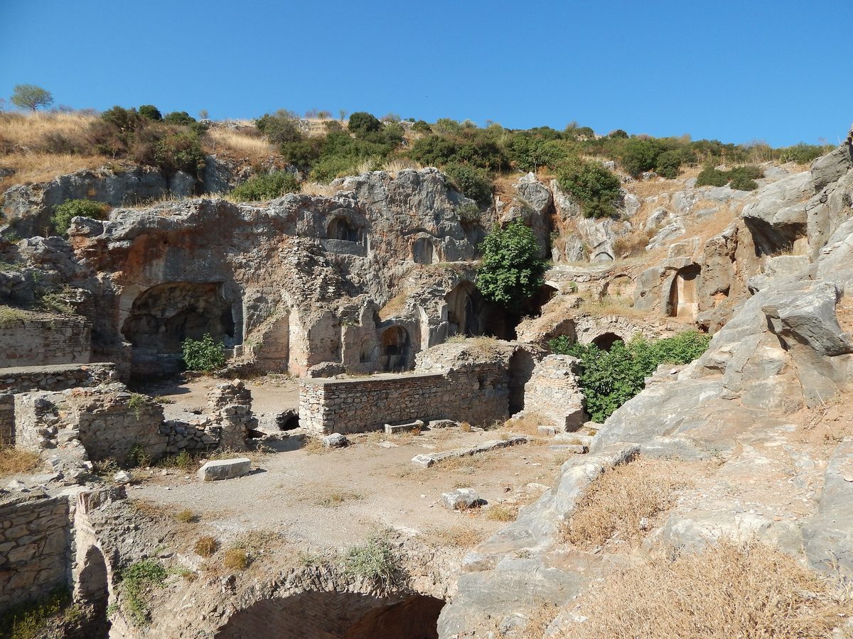 The ruins of the Seven Sleepers complex at Ephesus: brick and stone burial chambers below a limestone rock face cut with arched tomb niches, under a clear blue sky