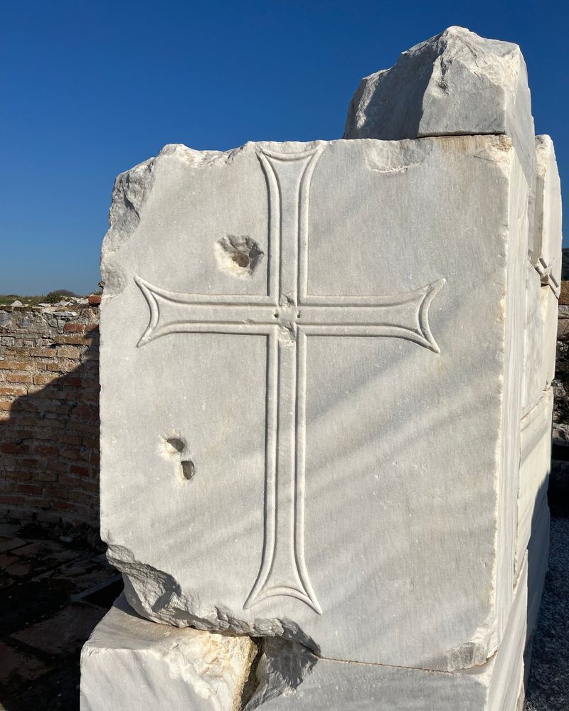 A tall cross with flared arms carved in outline on a white marble block among the ruins of Ephesus, against a deep blue sky