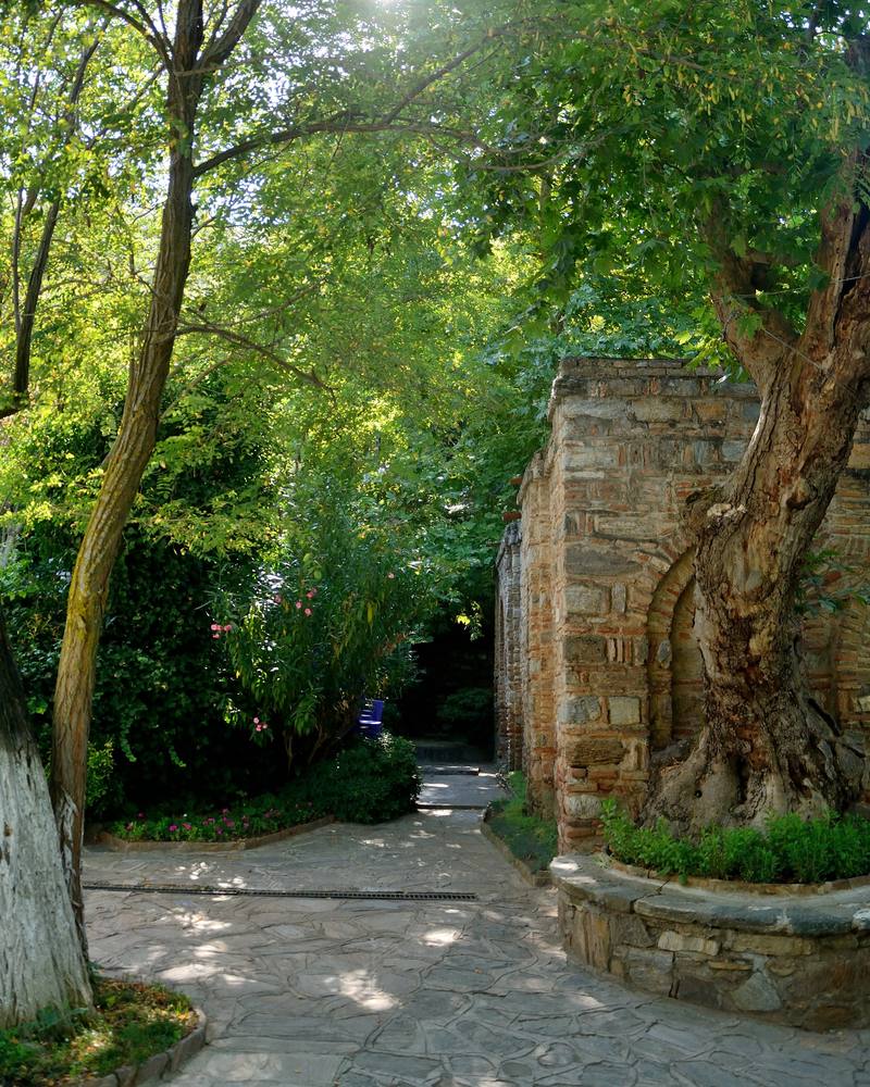 A shaded stone-paved garden path beside the brick-and-stone side wall of the House of Virgin Mary, under plane trees and green foliage