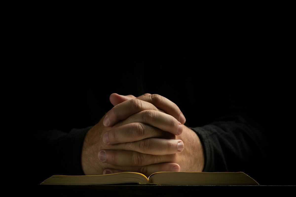 A person’s hands folded in prayer, resting on an open book with gilded page edges, against a dark background