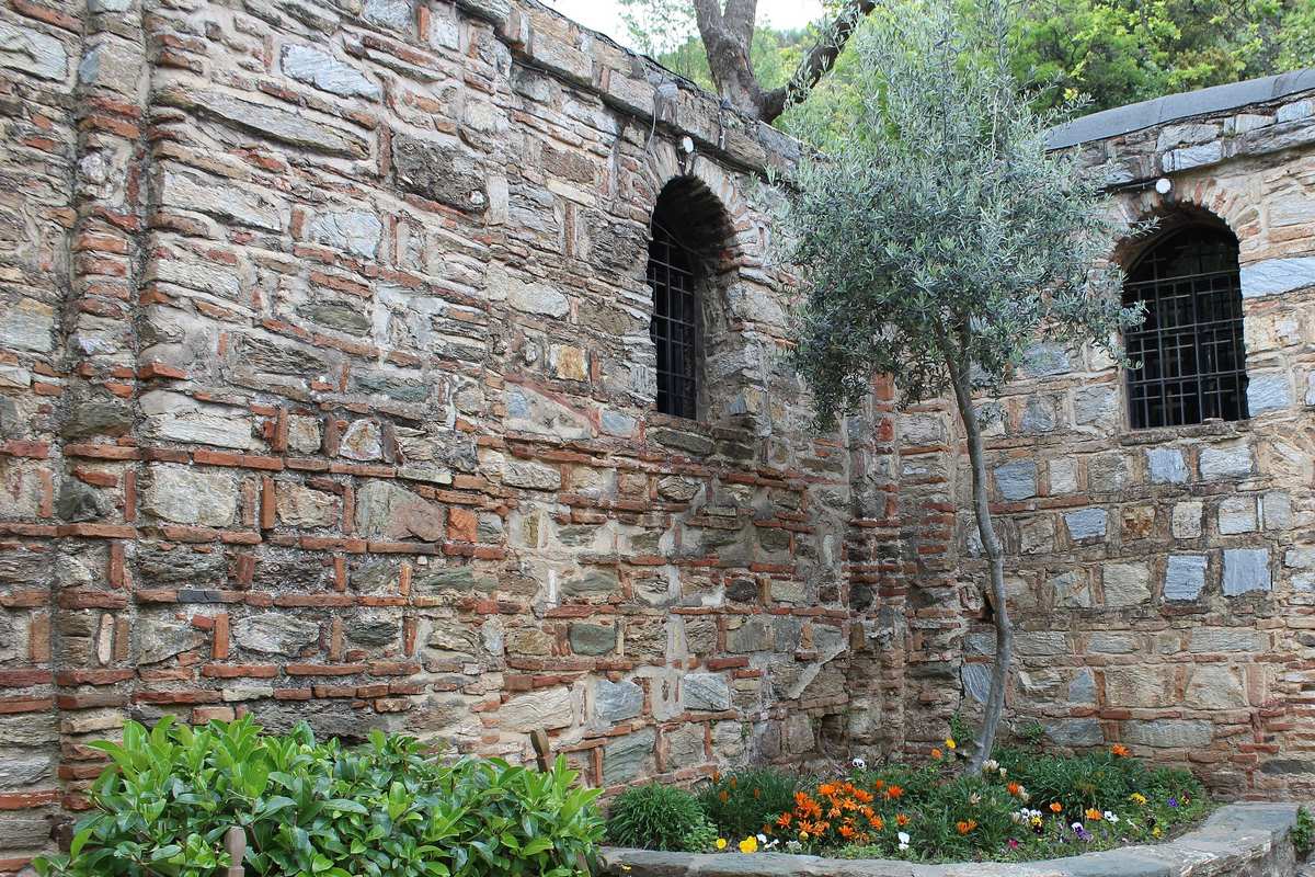 Gray stone walls laced with courses of red brick at the House of Virgin Mary, two small arched windows with iron bars, and a young olive tree in a flower bed where two walls meet