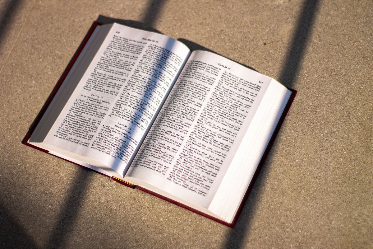An open Bible with a dark red cover lying on a stone floor, its pages crossed by bands of sunlight and shadow