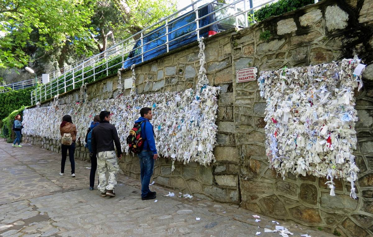 Young pilgrims standing on the stone path beside the wishing wall below the House of Virgin Mary, where thousands of white paper and cloth prayers are tied along the wall