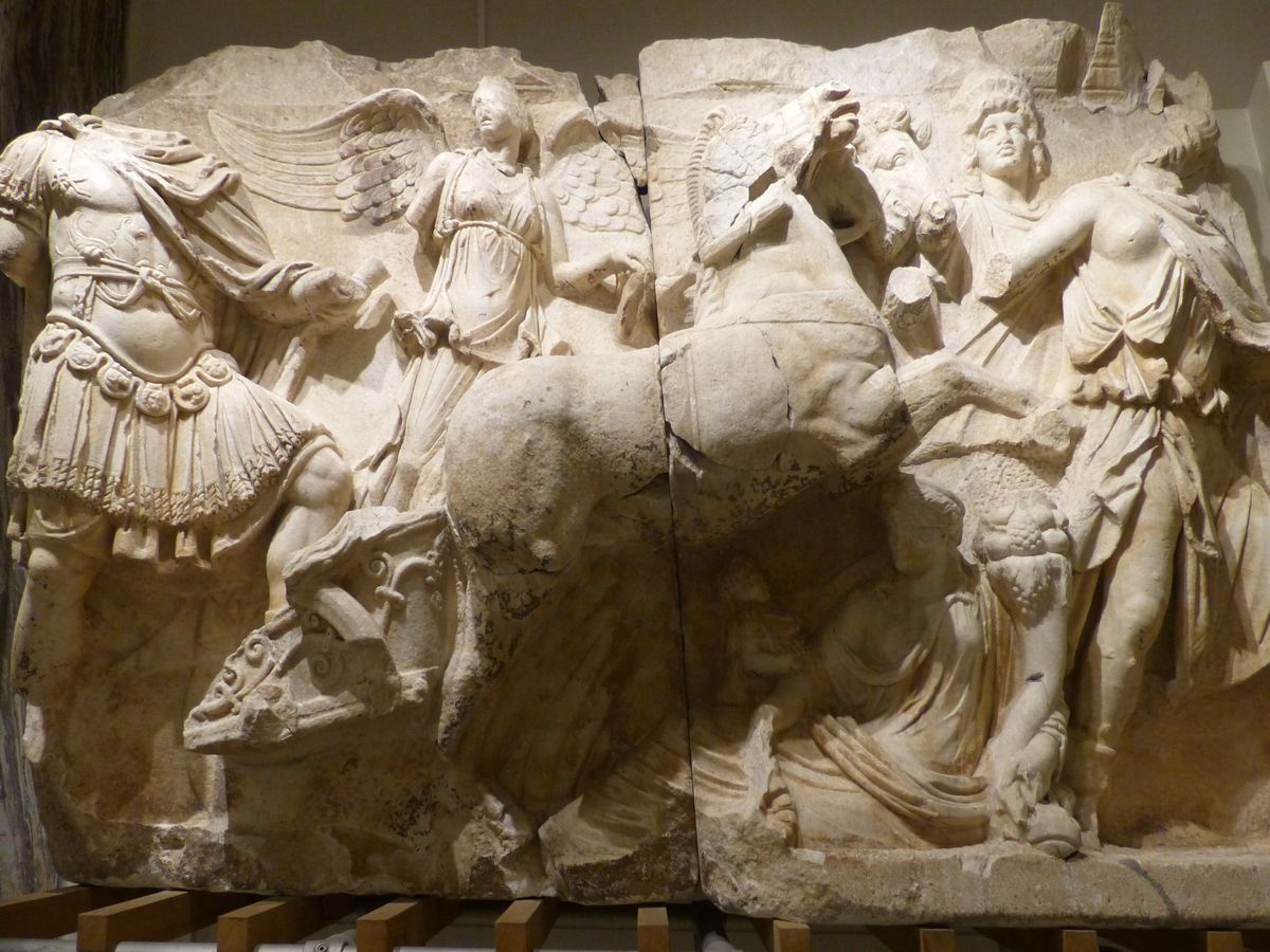 Marble relief of a headless Roman emperor in a molded breastplate, belt and fringed armored skirt, beside a winged Victory and a rearing horse