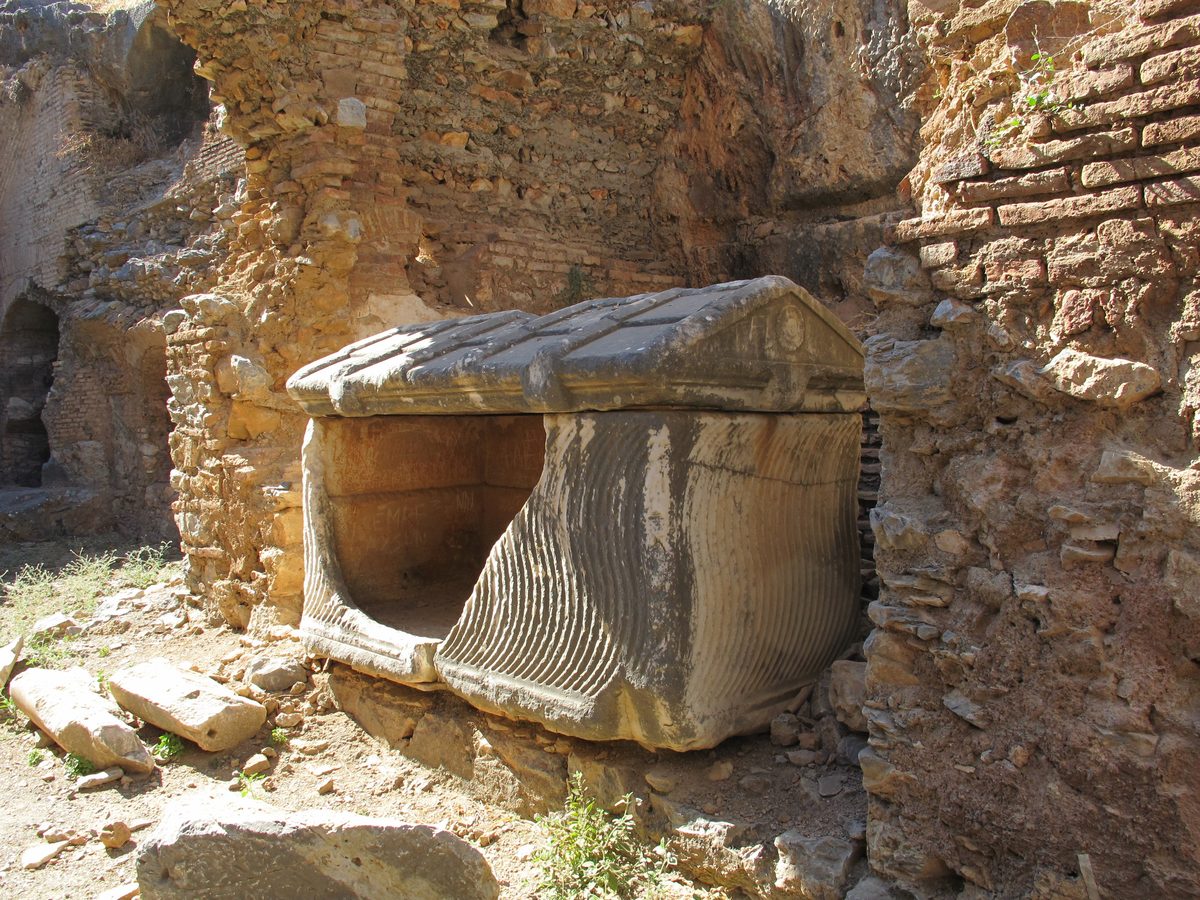 An open marble sarcophagus with a gabled lid and a fluted side, set among crumbling brick and stone walls at the Seven Sleepers cemetery in Ephesus