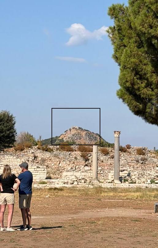 A rocky hilltop west of the ruins of Ephesus with a small stone tower on its summit, outlined by a black box, seen past a ruined wall and two standing columns